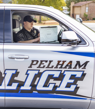 Image shows a Pelham Police officer in a police cruiser looking at a laptop installed in the vehicle
